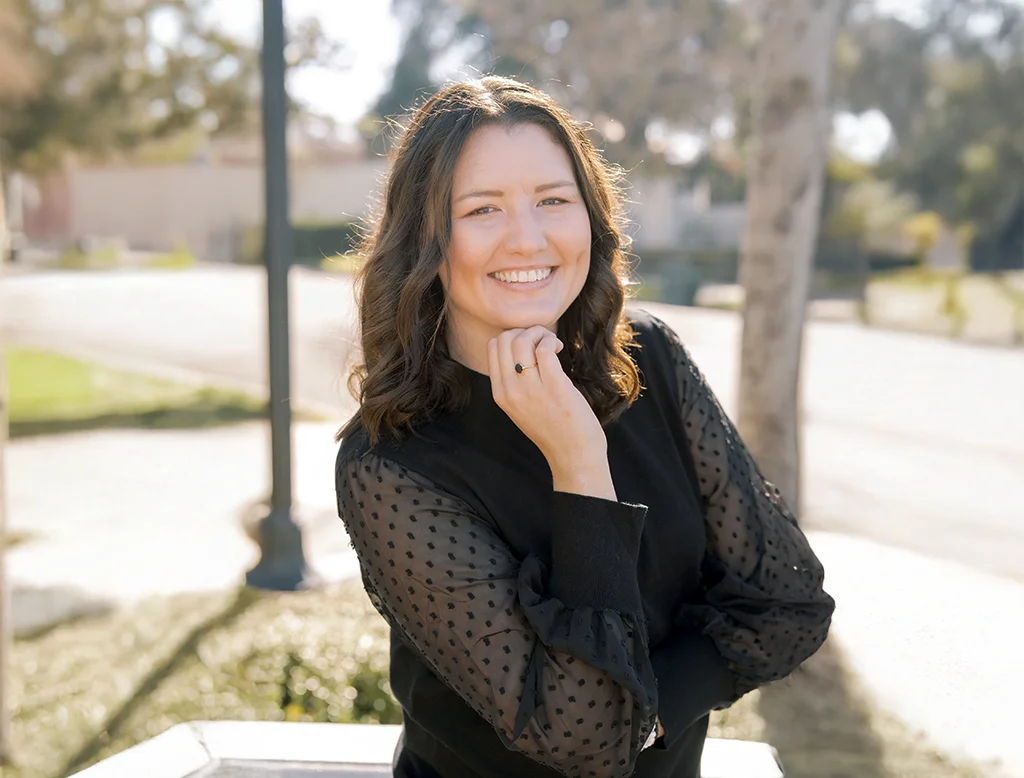 Woman with wavy brown hair smiling outdoors in a black top with sheer sleeves, posing confidently in natural light.